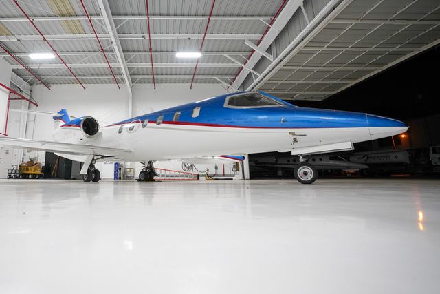 Blue and white business jet parked in a clean, bright hangar, reflected in the white floor.