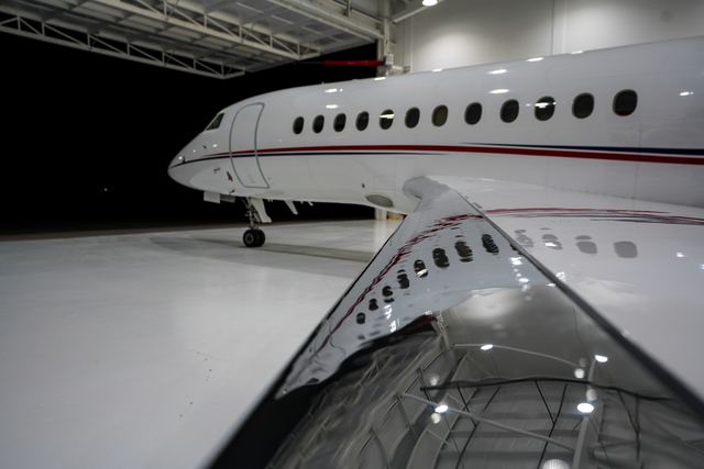 White business jet in a bright hangar, the hangar lights reflected in the polished top of its wing.