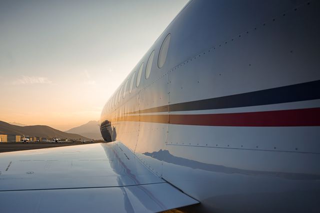 Glossy white business jet on the ramp at sunset, its navy and red cheatline running along the fuselage, with mountains reflected in the wing.